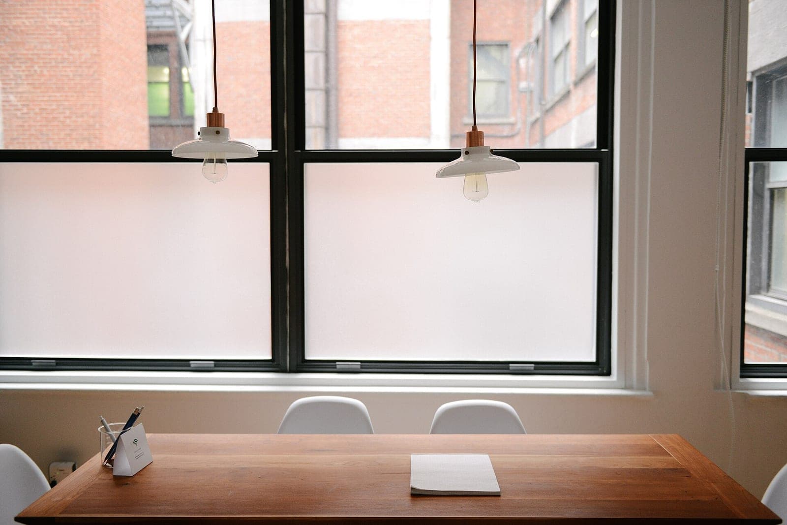 A wooden table under two pendant lamps in front of frosted office windows