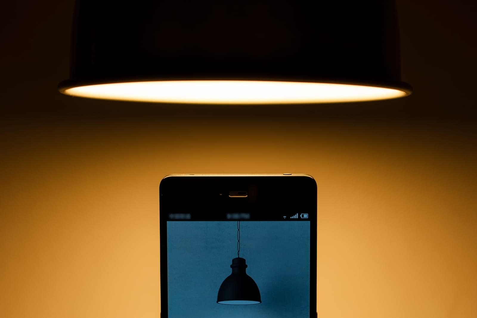 A smartphone stands upright beneath a hanging lamp that casts a warm orange glow