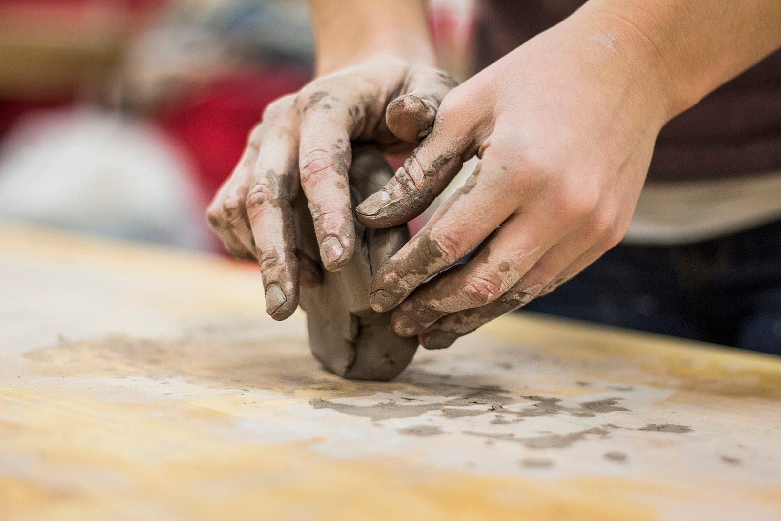 Two clay-covered hands pressing a lump of clay on a wooden worktable