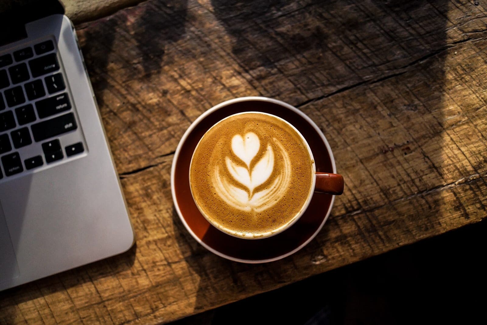 A latte with leaf-shaped foam art in a brown cup on a rough wooden table, beside the corner of a laptop keyboard