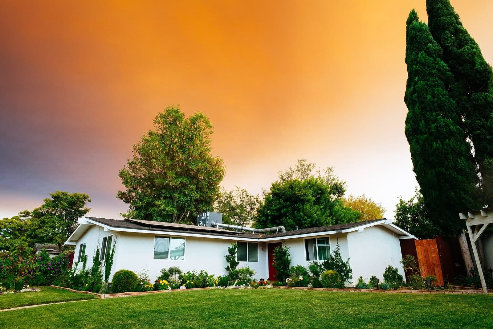 A white one-story house with a green lawn, flower beds and tall trees under an orange evening sky