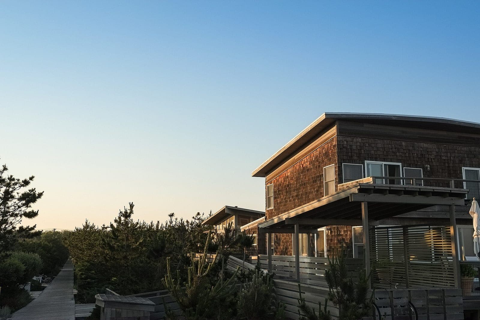 A wood-shingled house with a covered deck catches the evening sun beside a boardwalk path
