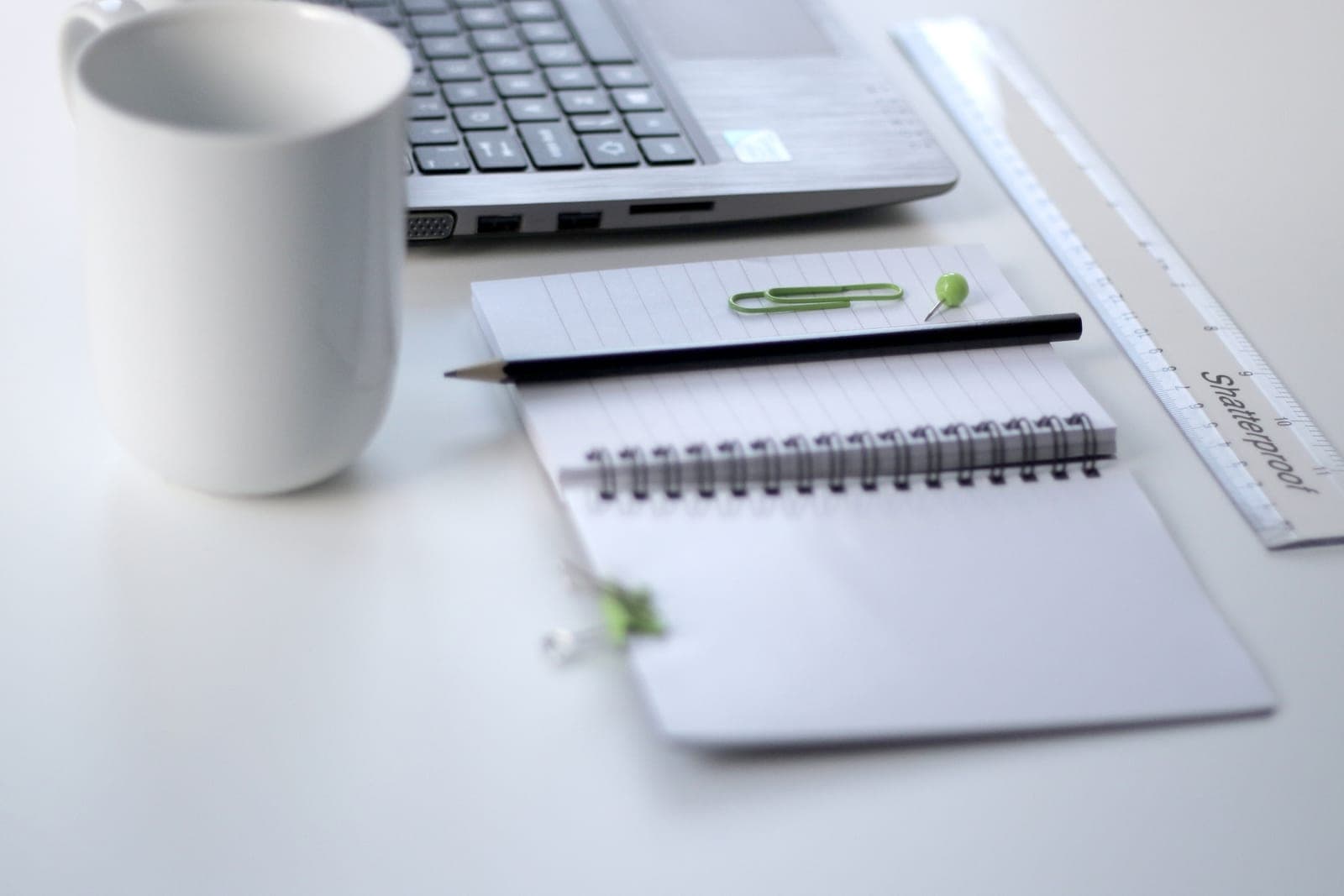 A laptop, an open spiral notebook with a pencil, a ruler and a white mug on a white desk