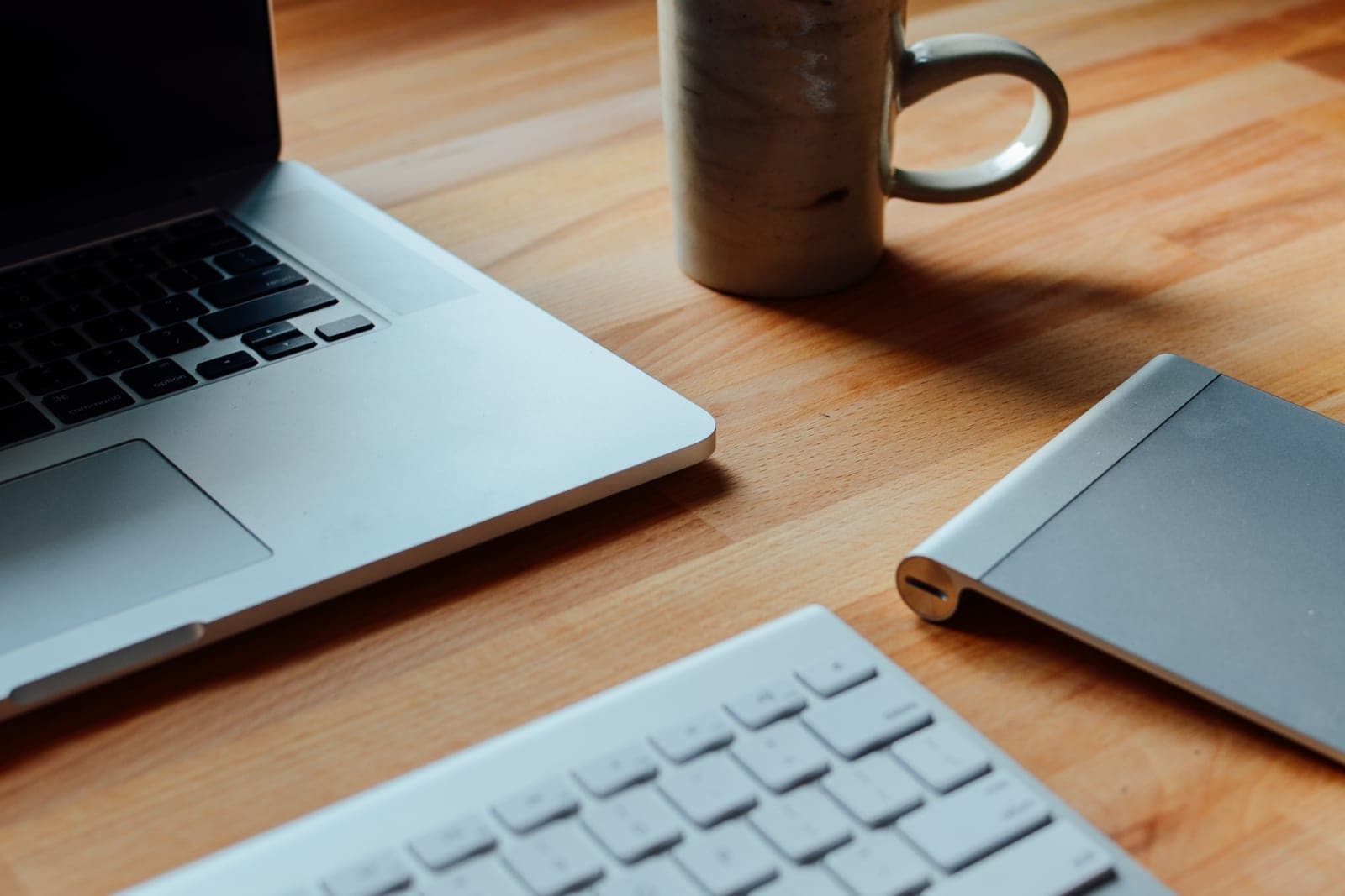 A laptop, a slim keyboard, a trackpad and a coffee mug on a wooden table
