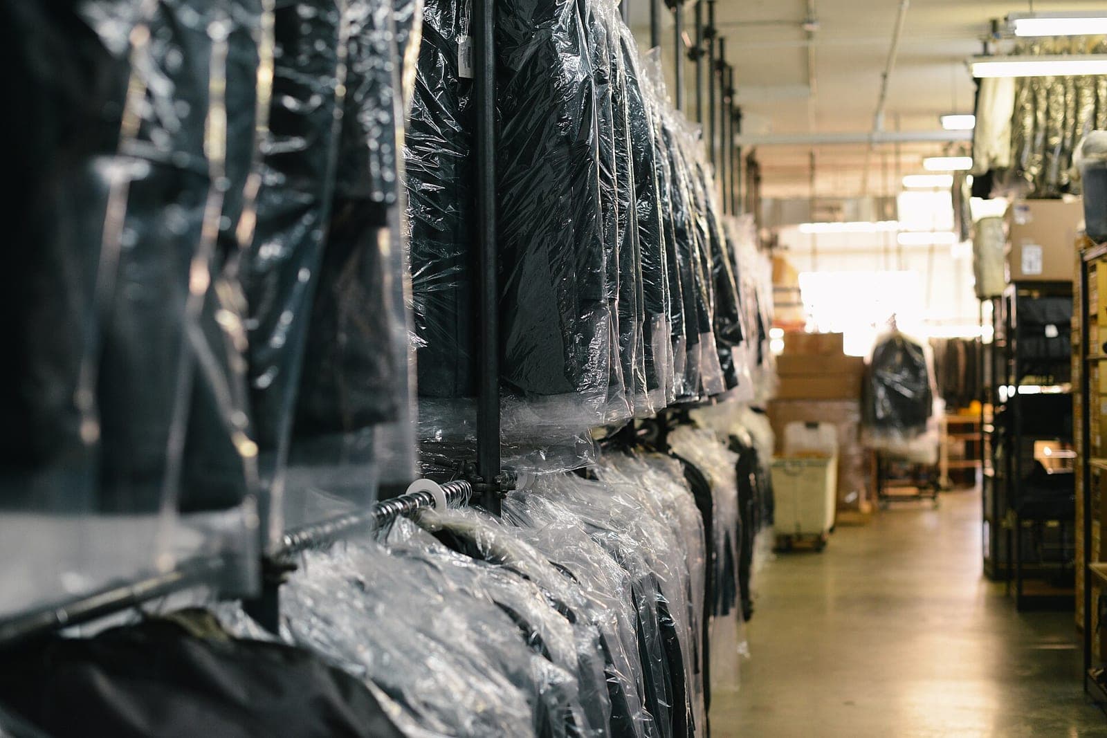 Rows of clothes in clear plastic bags hang on a long rail in a bright dry cleaning plant