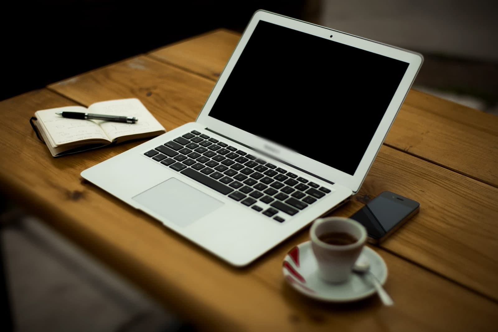 An open laptop, a notebook with a pen, a phone and a cup of coffee on a wooden table