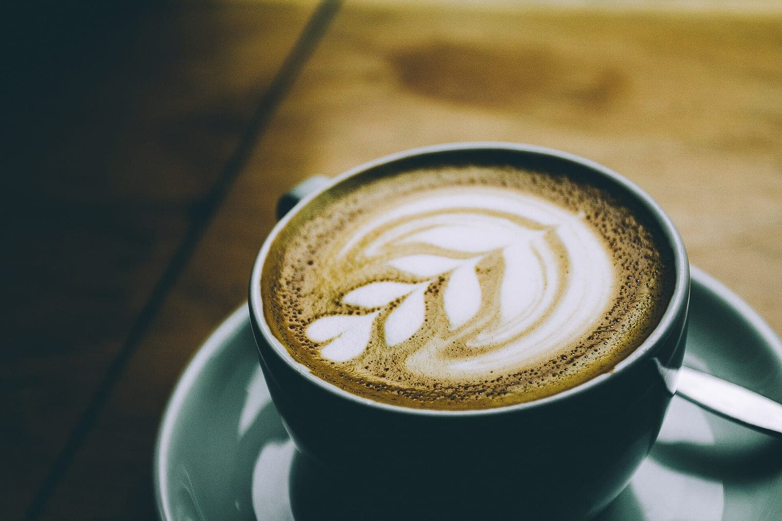 A cup of latte with leaf-shaped foam art on a saucer on a wooden table