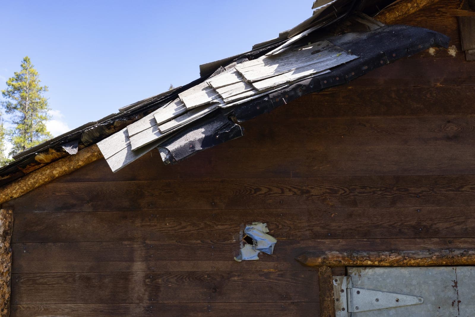 Loose and broken wood shingles hang over the edge of a roof above patched brown siding
