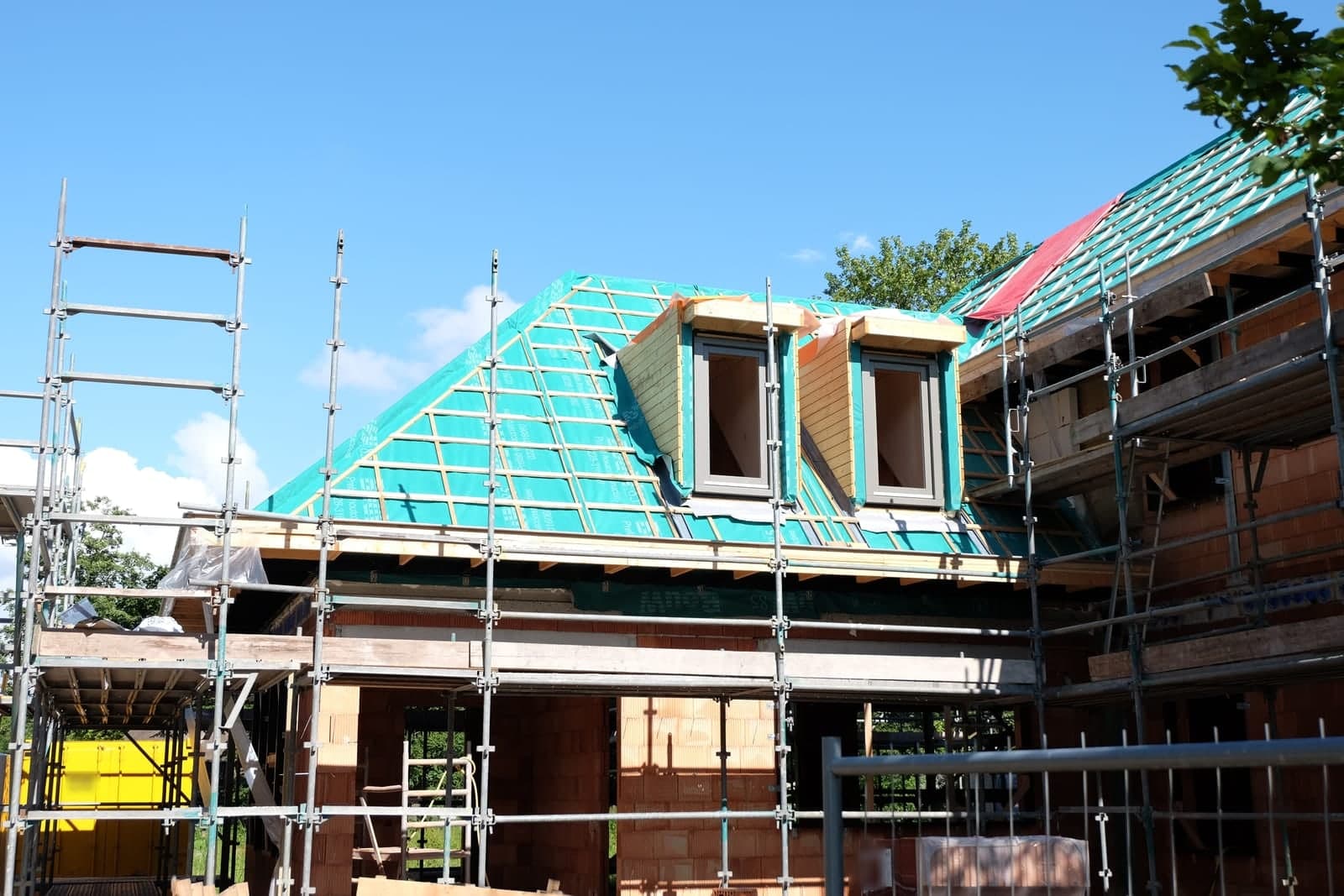 A new house under construction behind scaffolding, with dormer windows framed in a wrapped roof under a blue sky