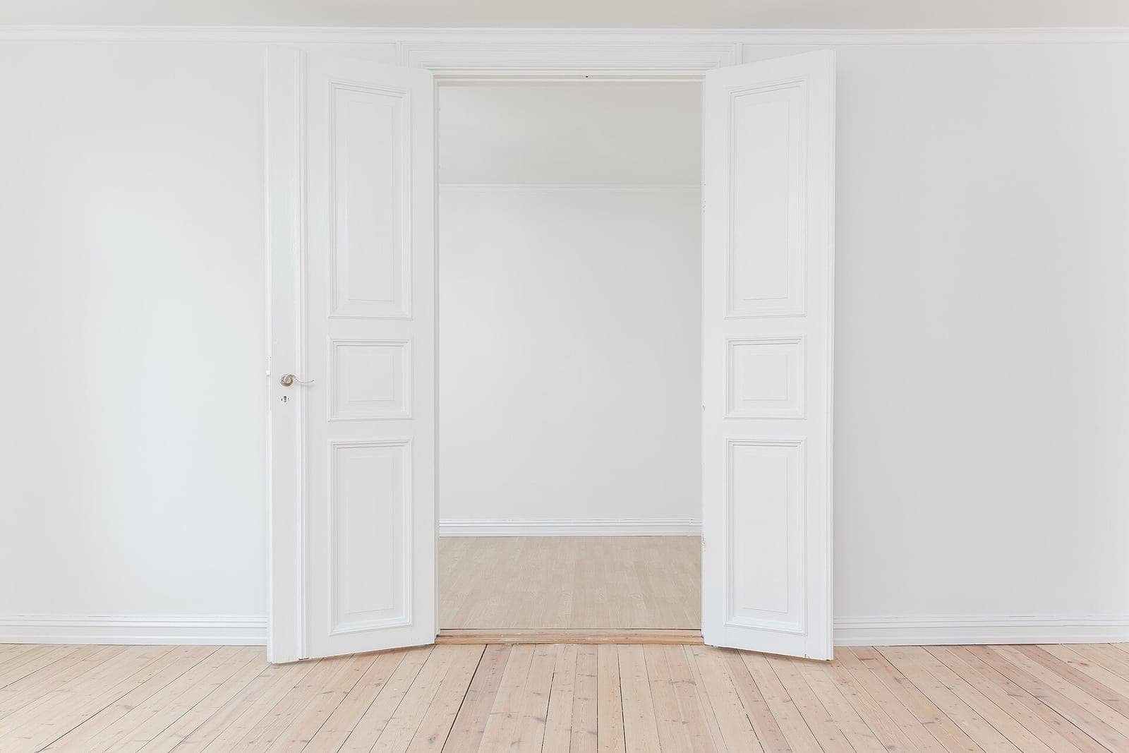 An empty room with white walls, open white double doors and a light wood floor