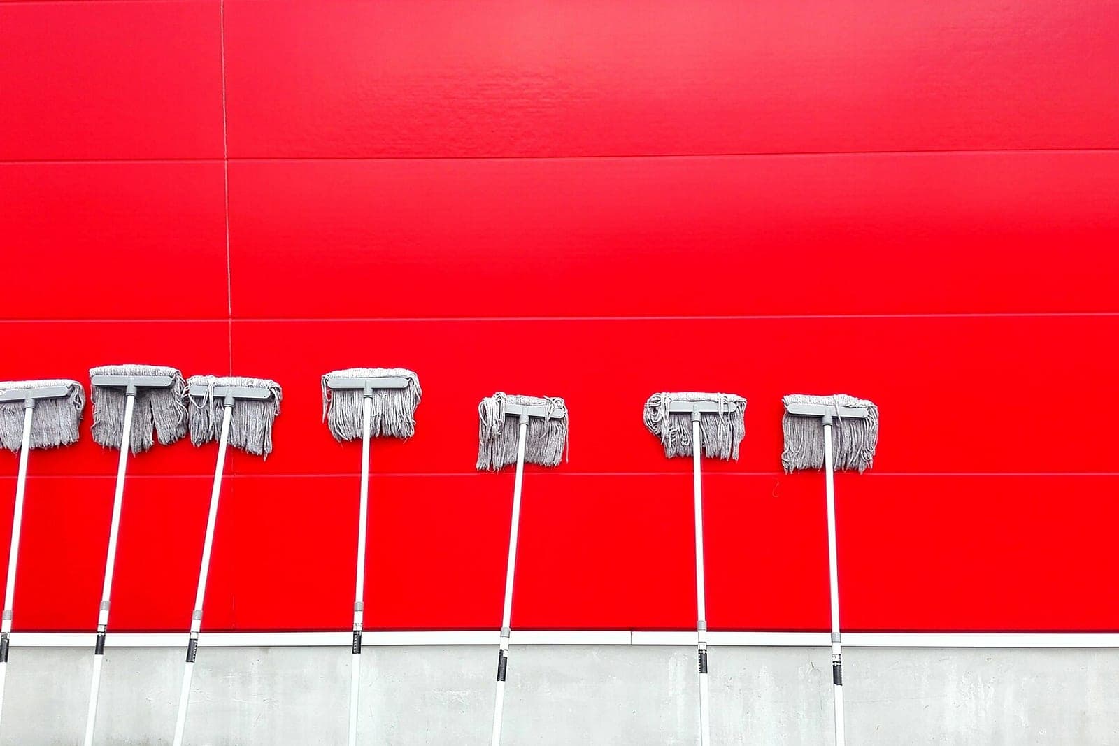 A row of gray mops stands head-up against a bright red wall