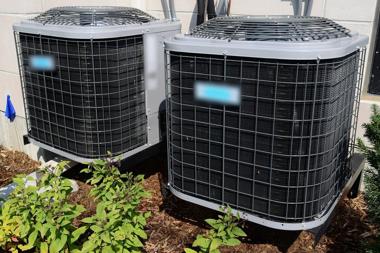 Two outdoor air conditioner condenser units side by side in a garden bed