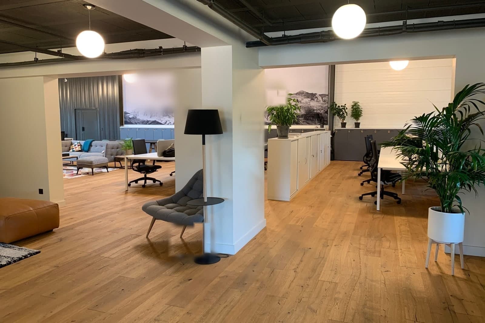 A bright open office with wood floors, hanging globe lights, rows of desks, plants and a lounge corner