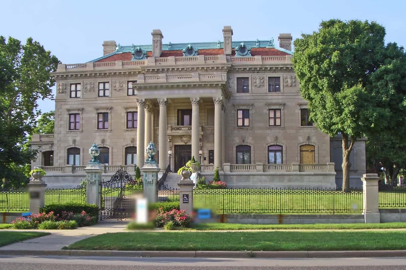 The Kansas City Museum's stone mansion stands behind iron gates and flower beds on a sunny day in Kansas City, Missouri