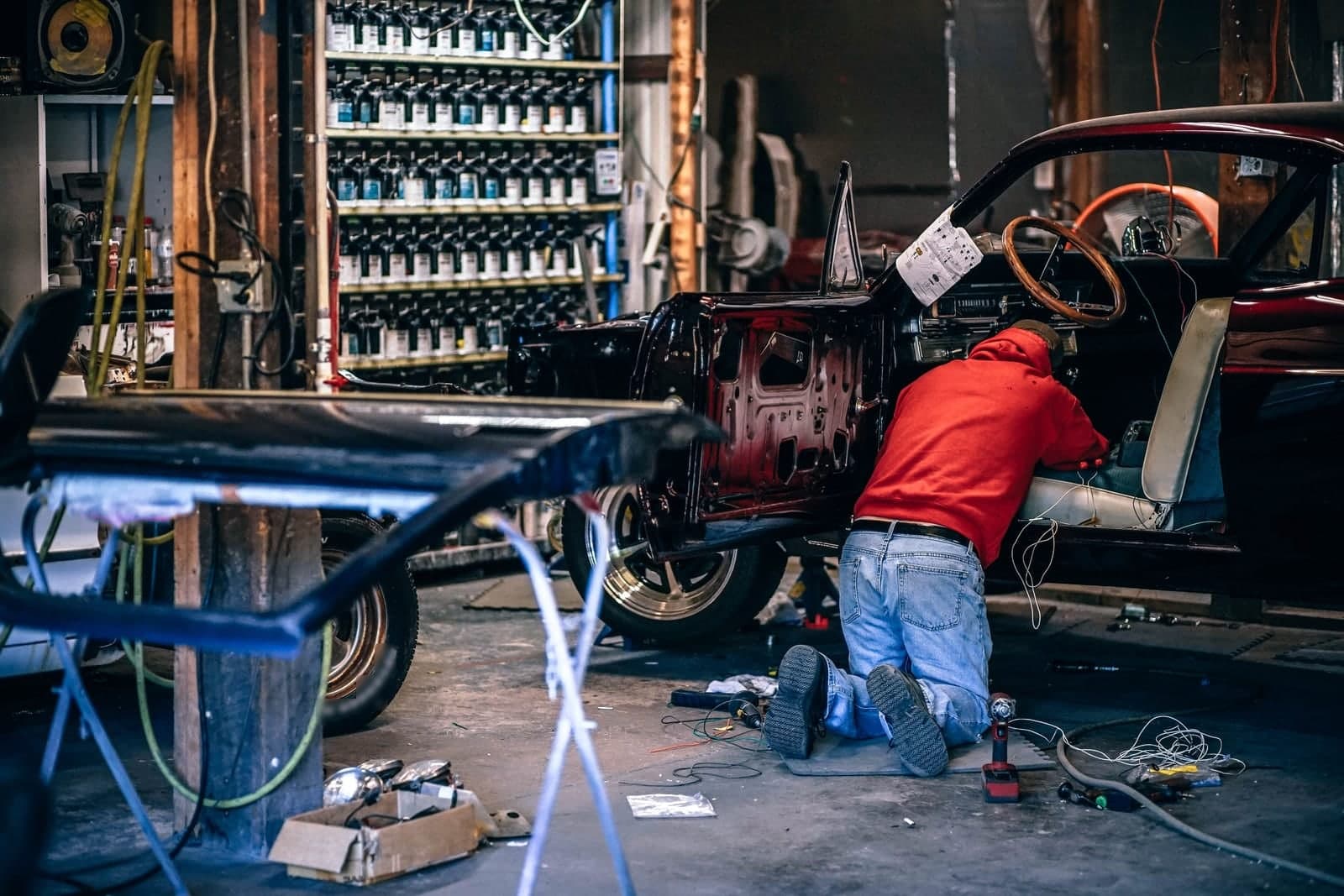 A mechanic, seen from behind, kneels to work under the dashboard of a red classic car in a busy repair shop