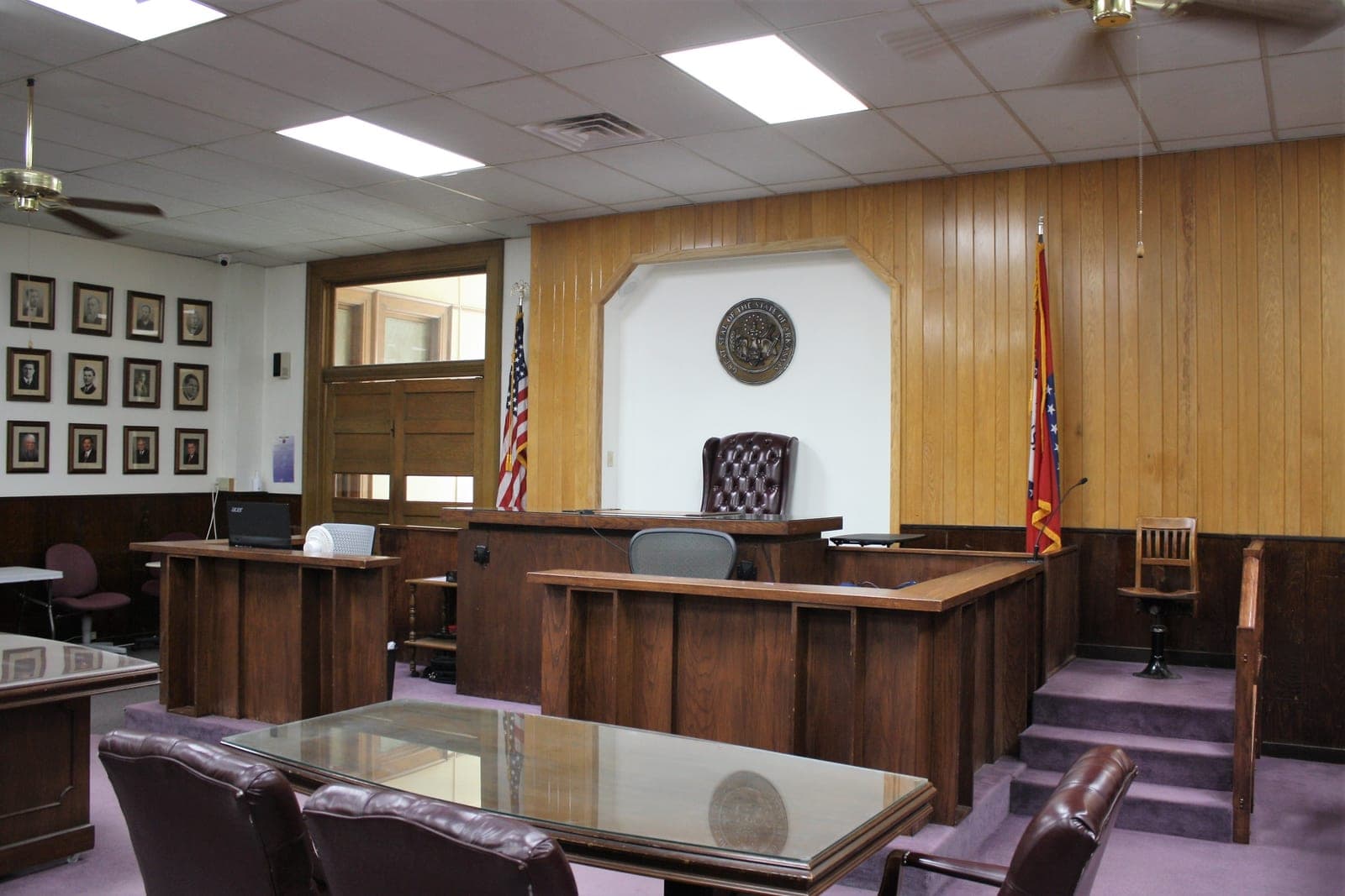 An empty small-town courtroom with a wood-paneled judge's bench, flags and counsel tables