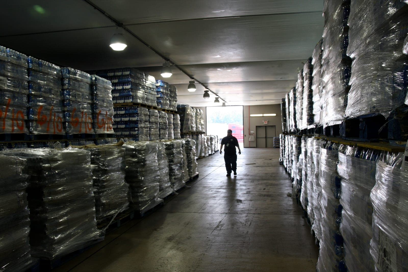 A worker walks down an aisle between tall rows of shrink-wrapped pallets in a warehouse