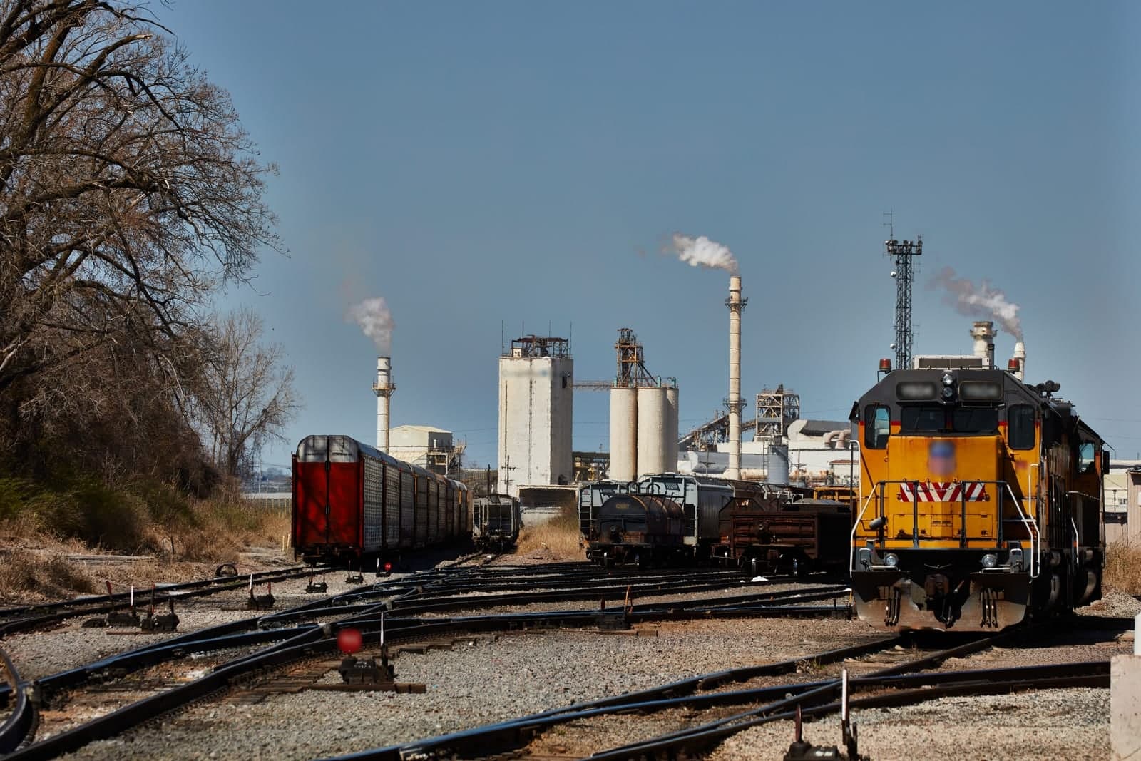 A railroad switchyard and industrial plant with tall silos and smokestacks under a blue sky in Kansas City, Kansas