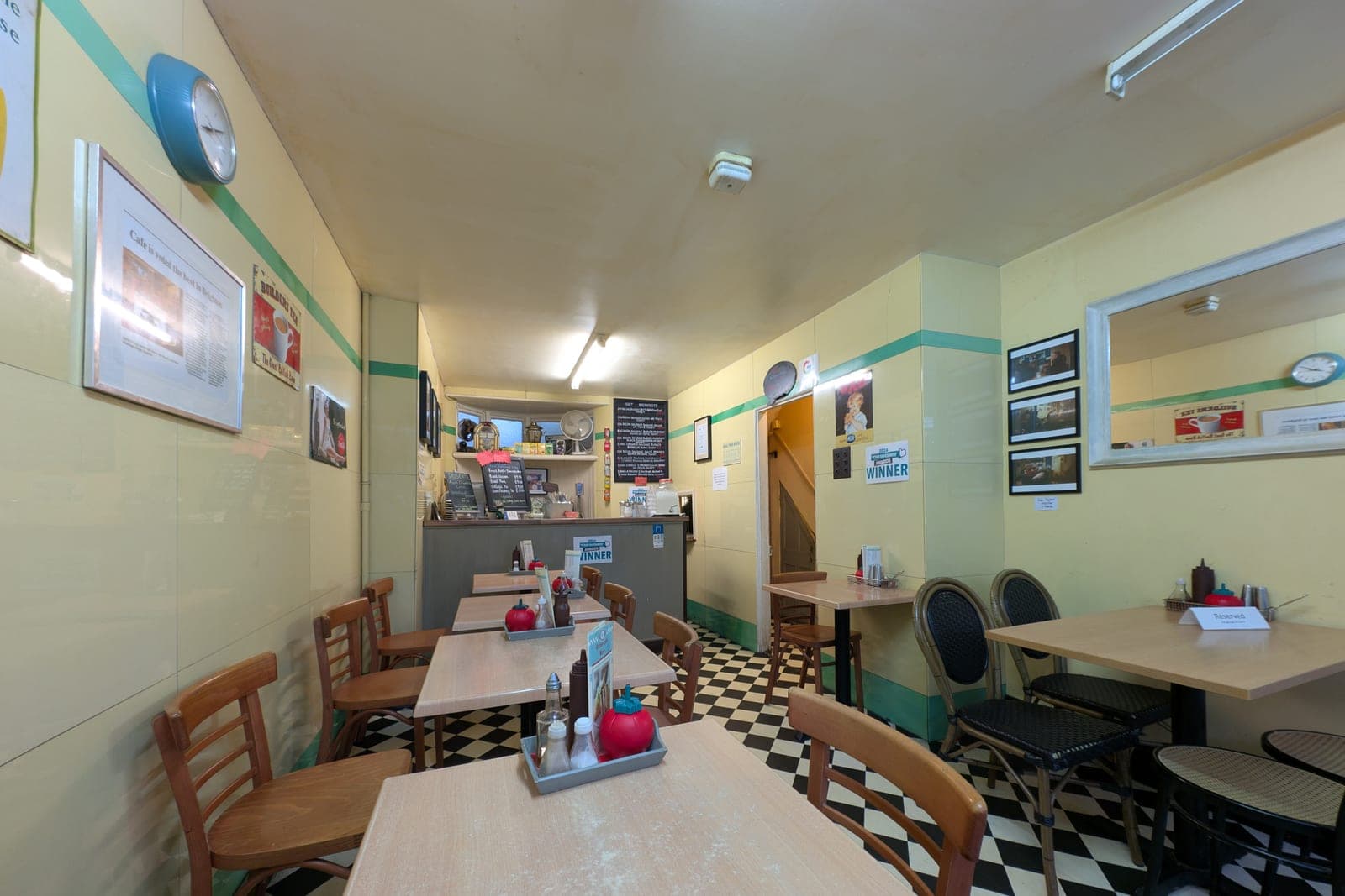 A small cafe with wooden tables and chairs, a checkered floor and a counter at the back