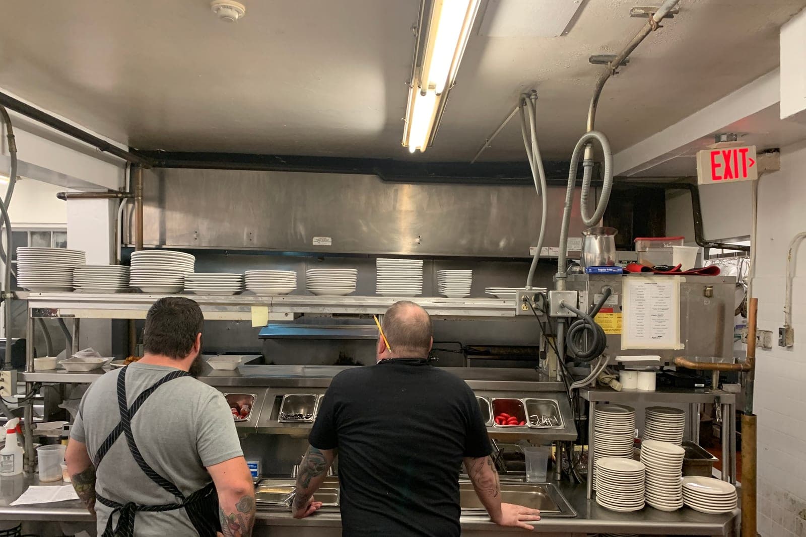 Two cooks stand at the expo station of a restaurant kitchen, with stacks of white plates on the shelf above