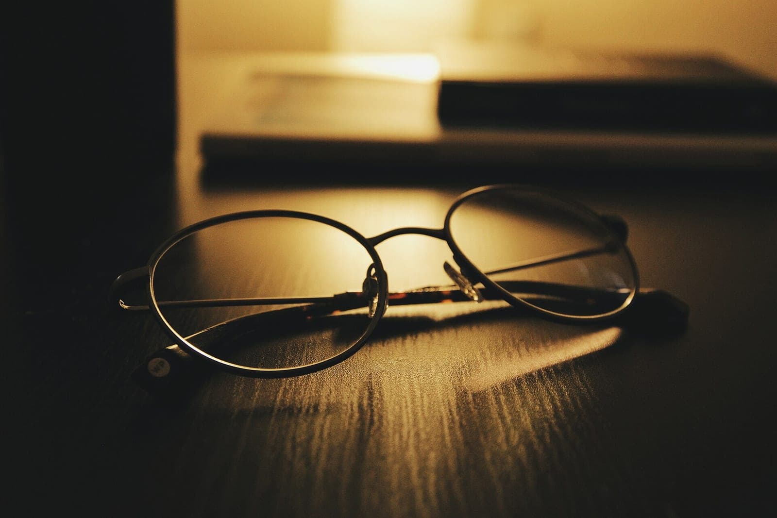 Round wire-frame glasses on a dark wooden desk in warm lamp light