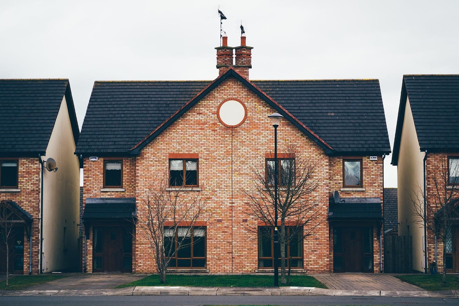 A brick semi-detached house with neighbors close on each side and bare young trees out front