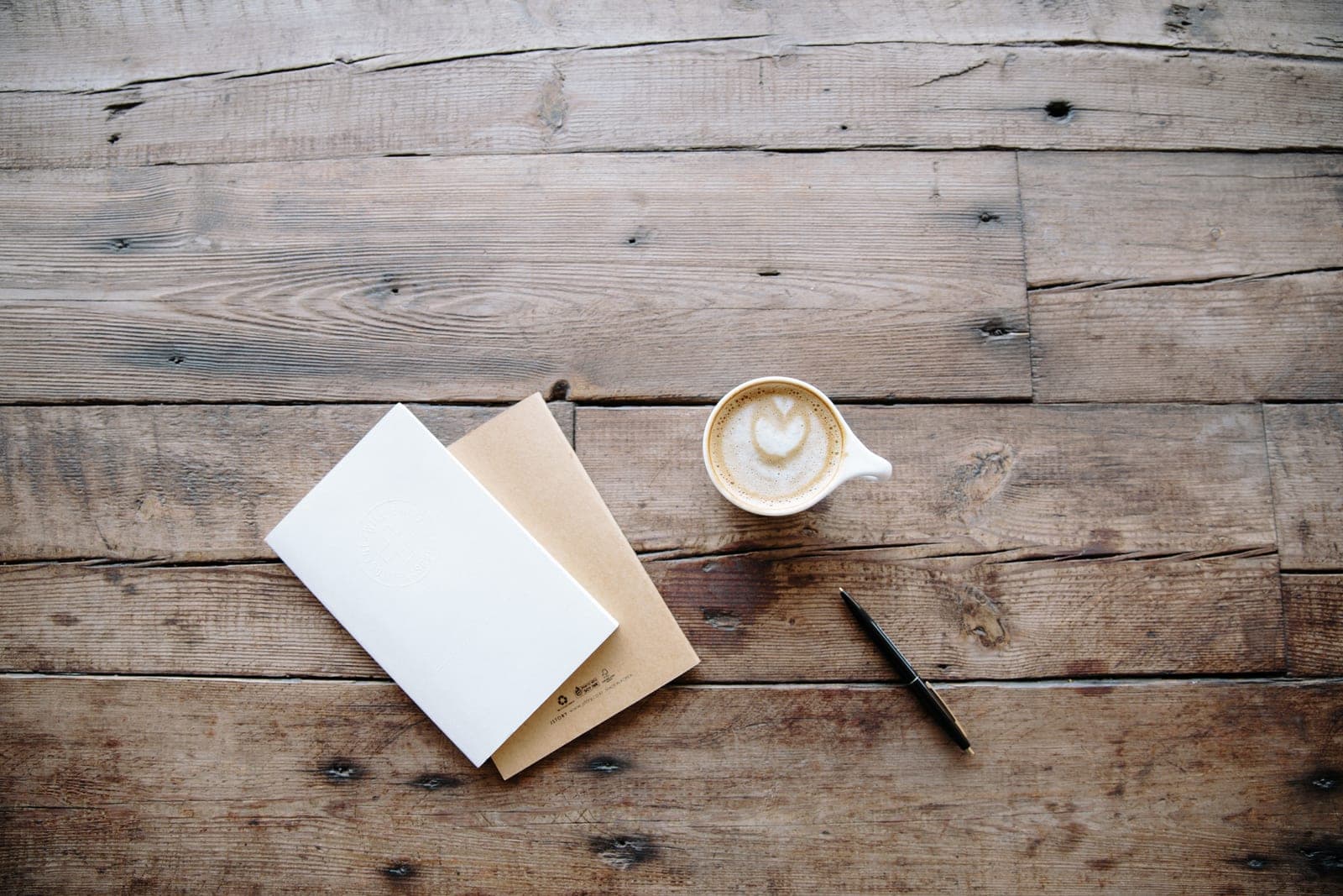 A white notebook, a tan notebook, a pen and a latte on a wooden table
