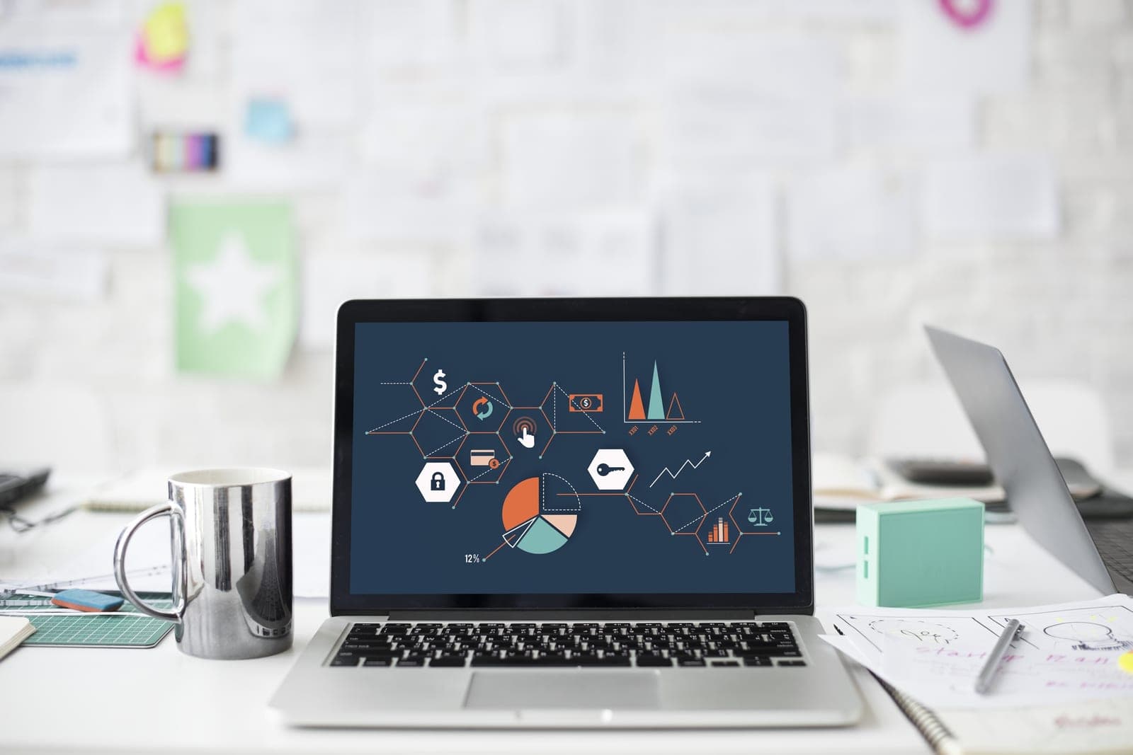 A laptop showing charts and simple icons on a white desk beside a metal mug and sketch papers