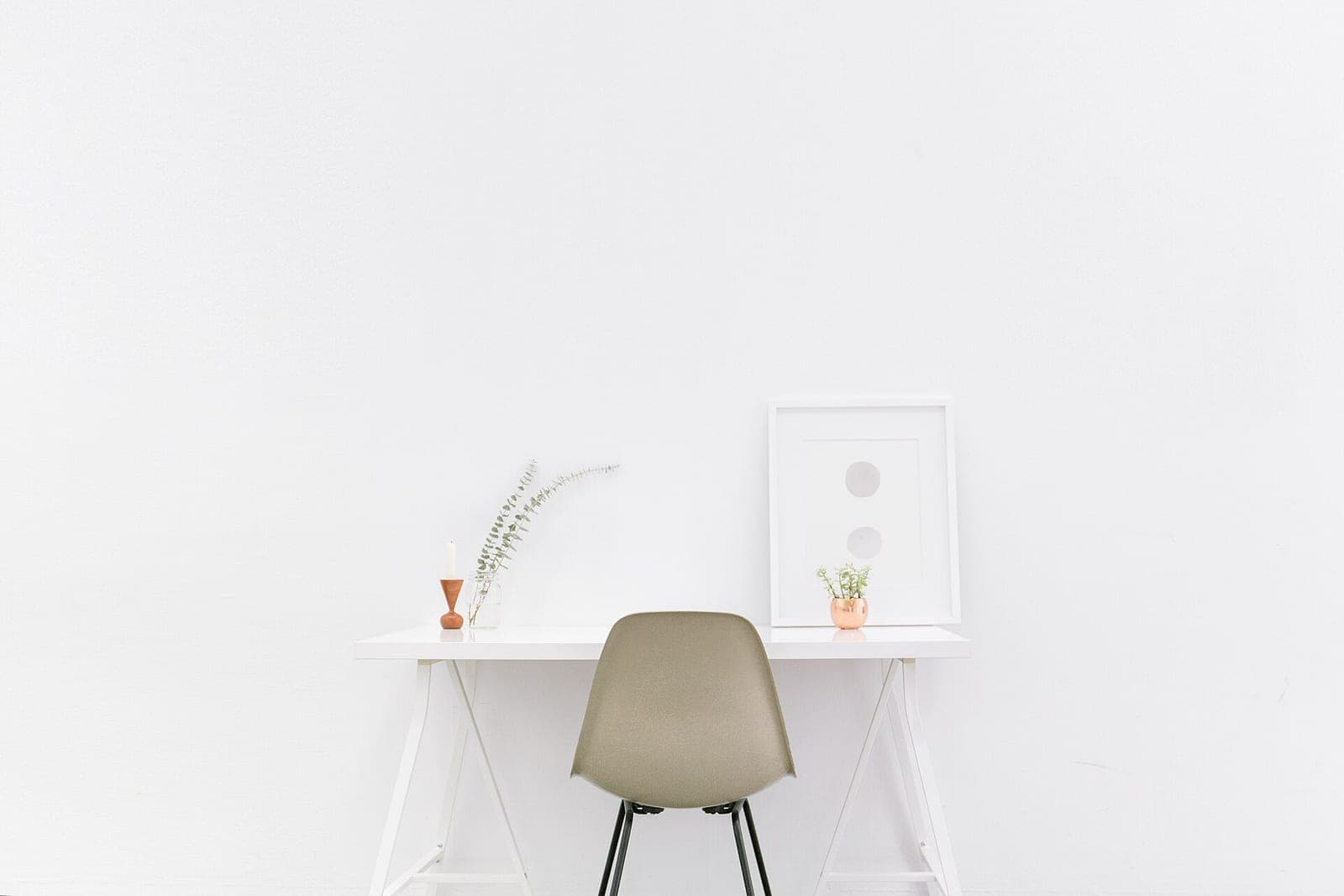 A white desk and a single grey chair against a plain white wall, with a small plant and a framed print