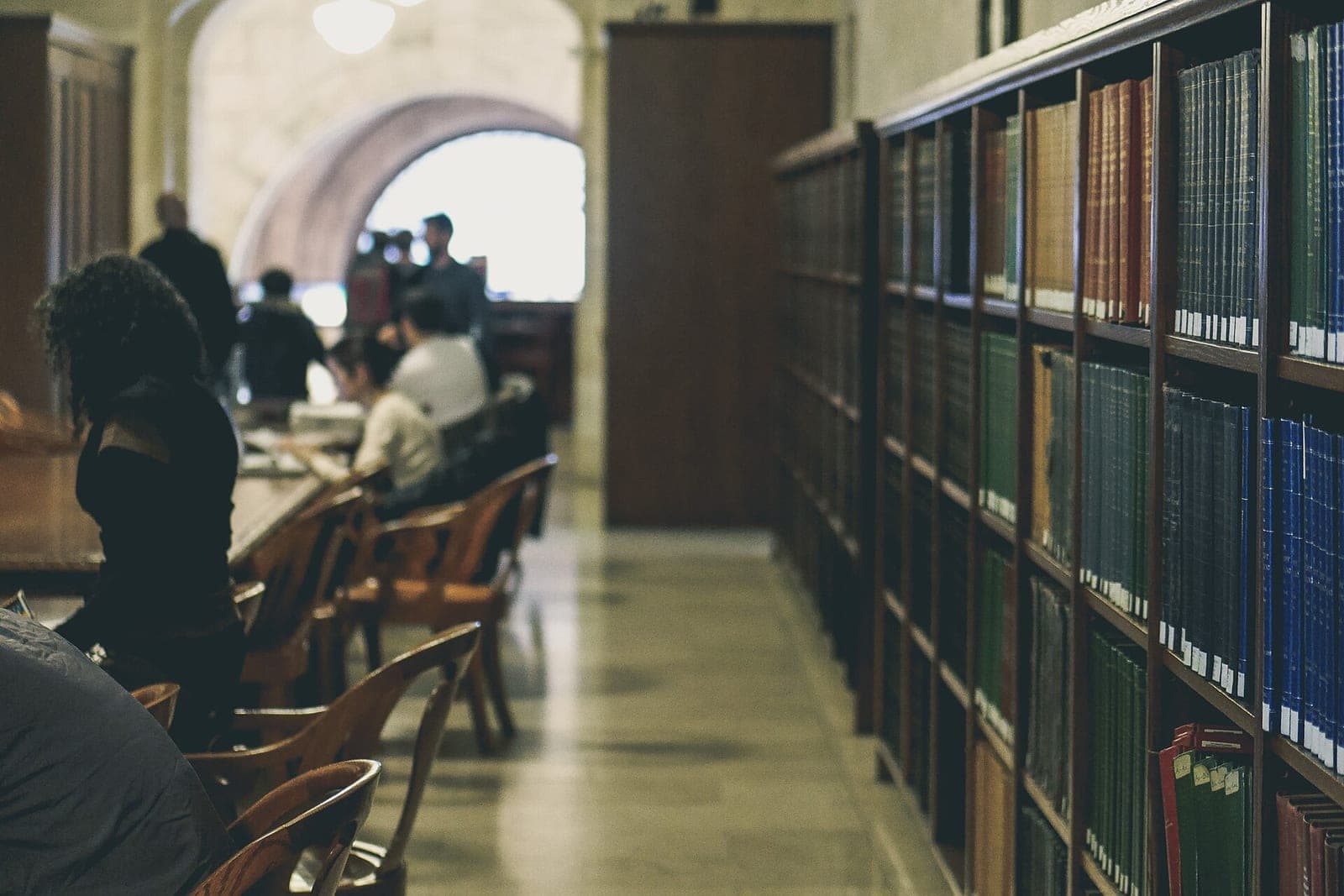 People reading at long wooden tables beside tall shelves of bound volumes