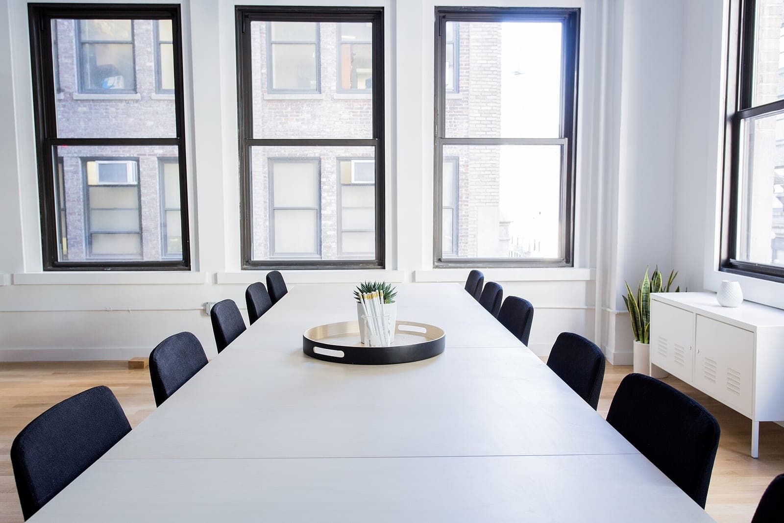 A long white meeting table with black chairs in a bright room with tall windows