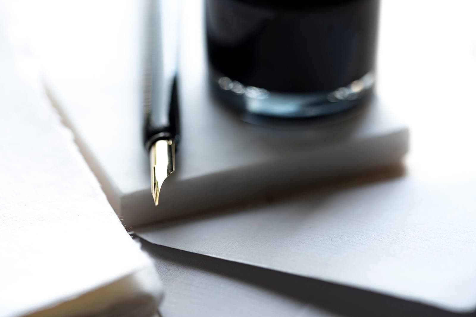 A pen with a gold nib rests on sheets of blank white paper beside a bottle of ink