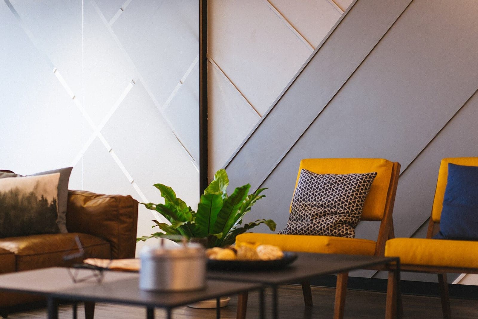 A lounge with two mustard-yellow armchairs, a brown leather sofa, a low table and a leafy plant against a gray paneled wall
