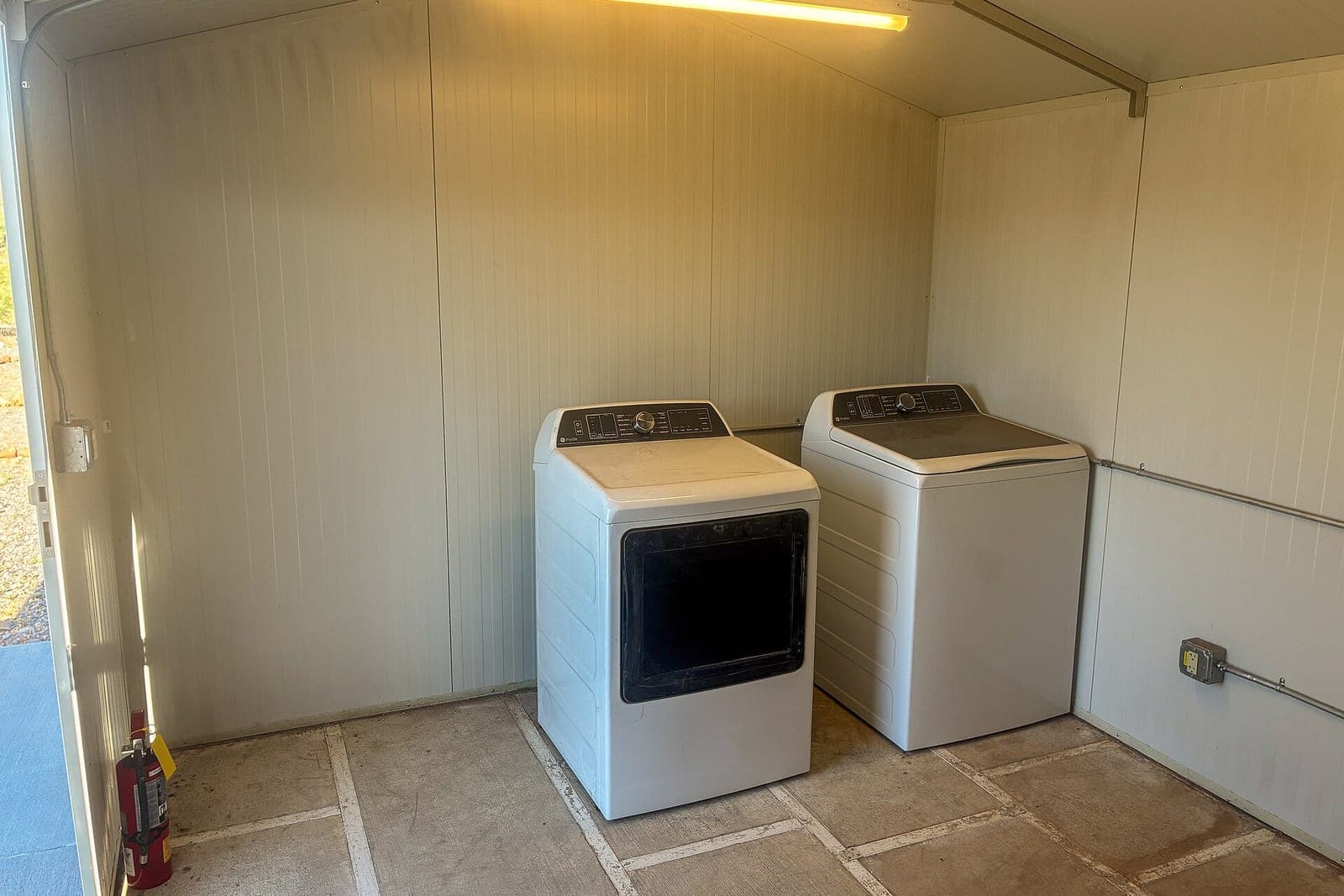 A white dryer and a white washer side by side in a small, plain laundry room under a ceiling light