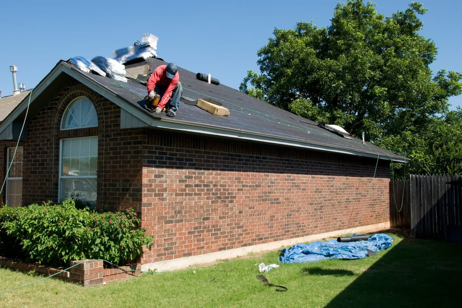 A roofer in a red shirt kneels on the roof of a one-story brick house, with shingle bundles stacked near the peak and a tarp on the lawn