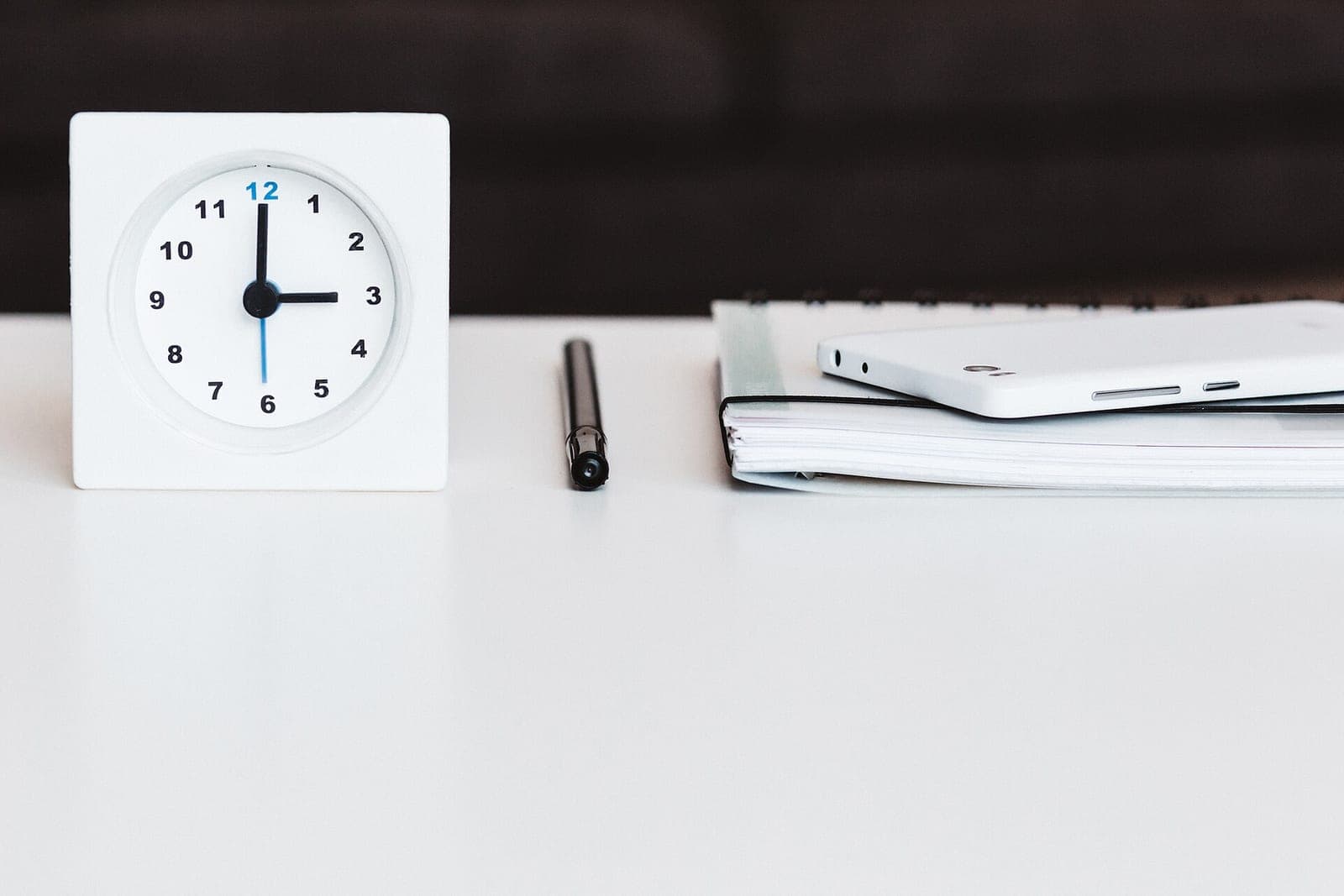 A white desk clock showing three o'clock beside a black pen and a white phone on a spiral notebook