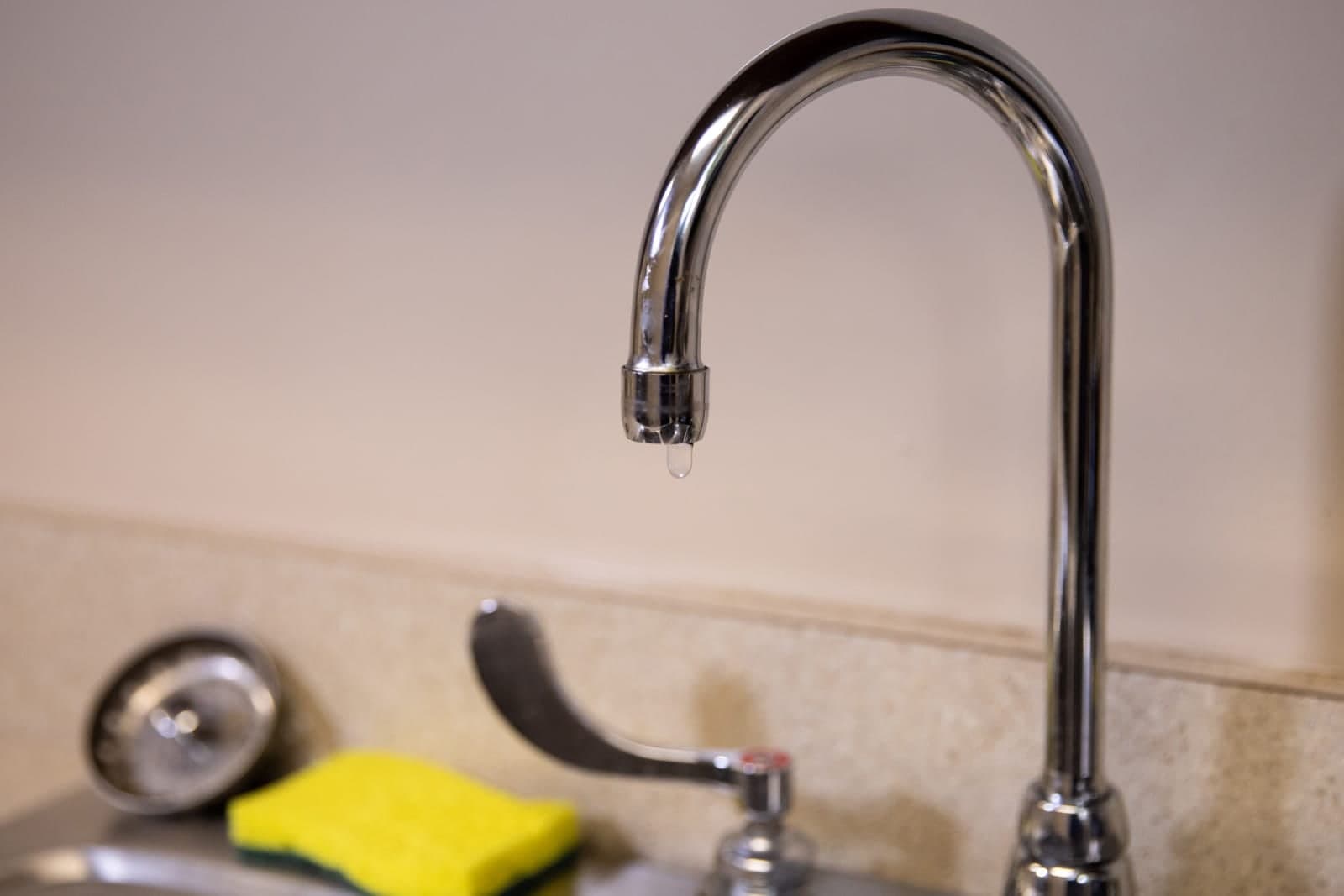 A chrome faucet drips a drop of water over a sink in a bright room