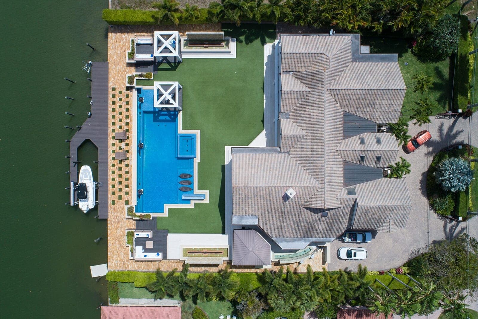Overhead view of a long blue pool, a lawn and a tiled-roof house beside a canal with a boat dock