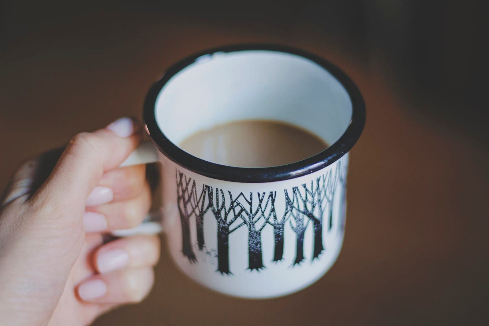 A hand holding a white enamel mug with a black tree pattern, half full of coffee with milk