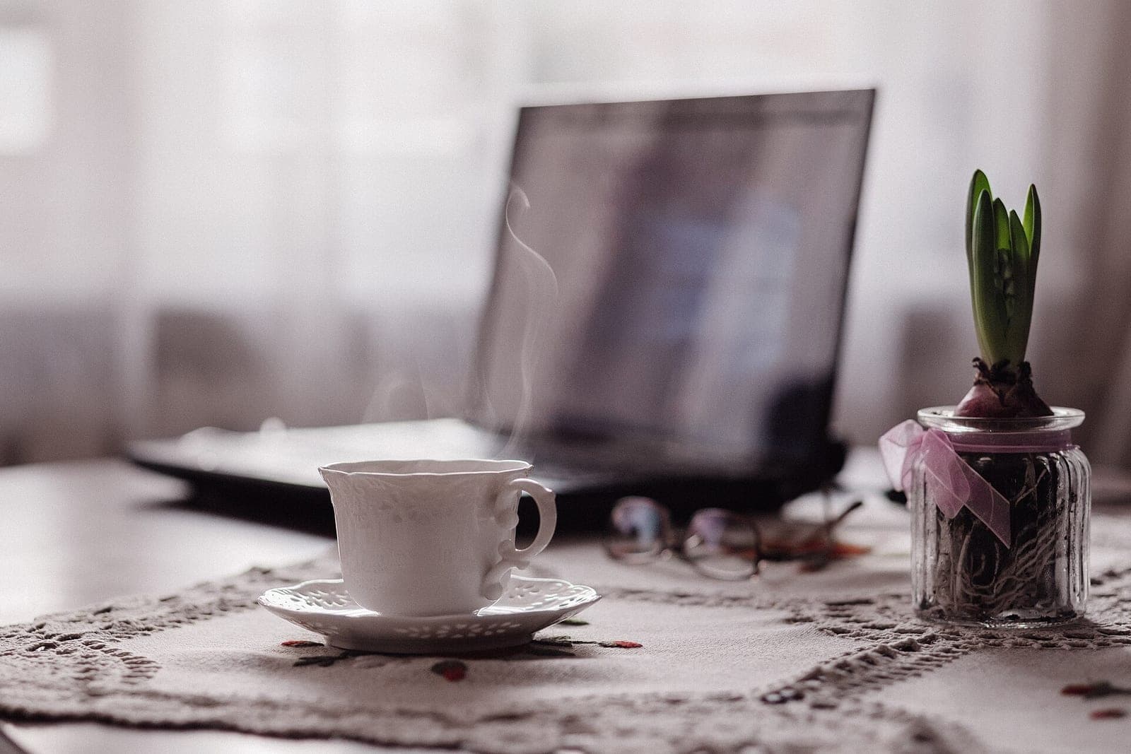 A steaming white teacup on a lace table runner, with reading glasses, a hyacinth bulb in a glass jar and a laptop behind