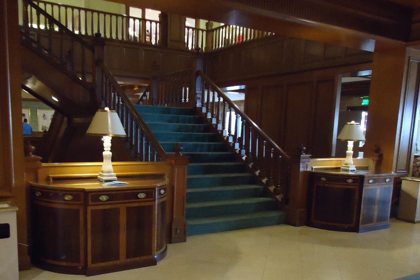 Two curved wooden desks with table lamps stand at the foot of a carpeted staircase in a quiet lobby