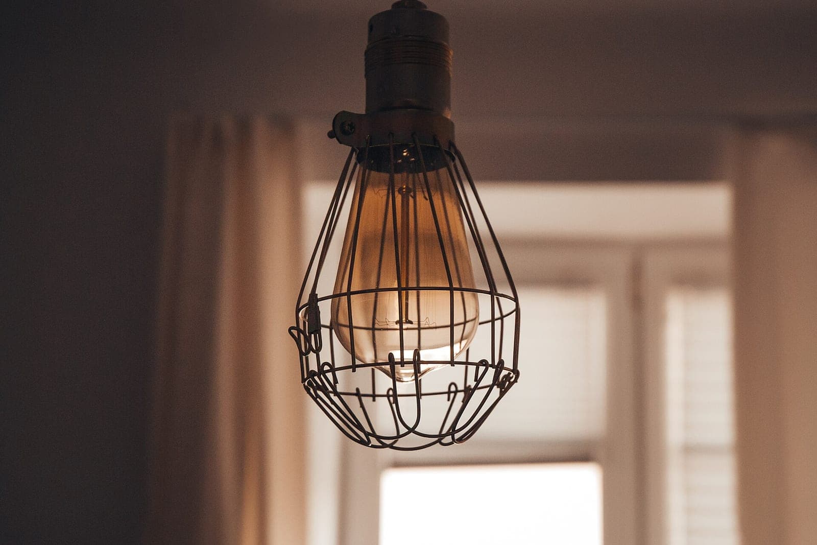 A bare light bulb glows inside a black wire cage in front of a bright window