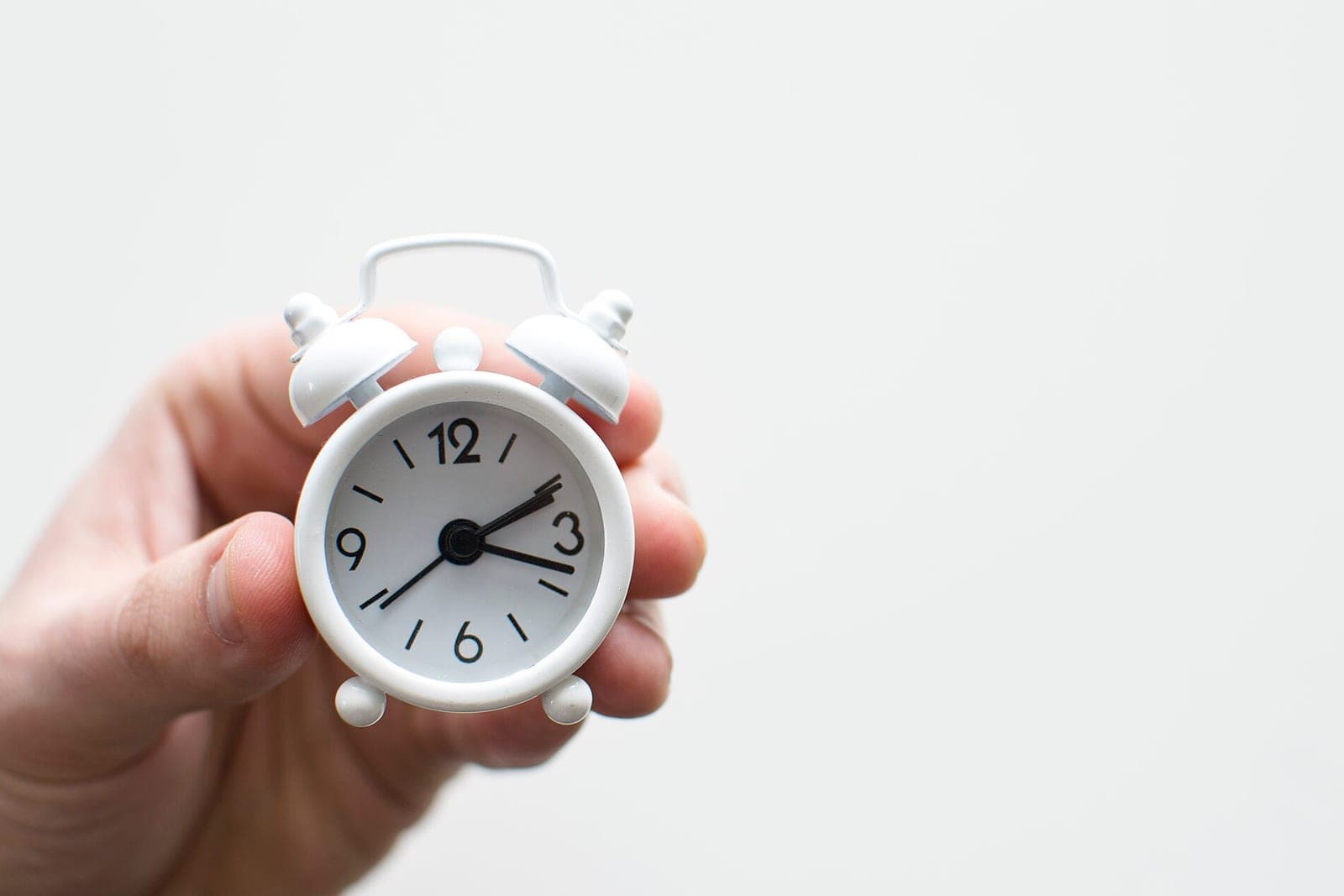 A hand holding a small white alarm clock against a plain white wall