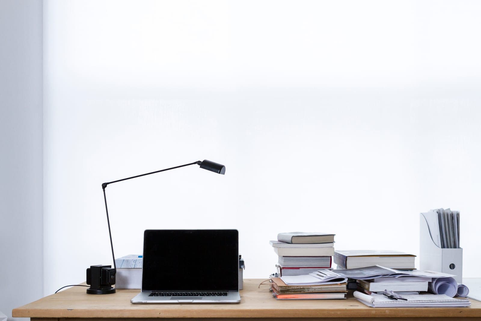 An open laptop on a wooden desk beside a desk lamp and stacks of books and papers