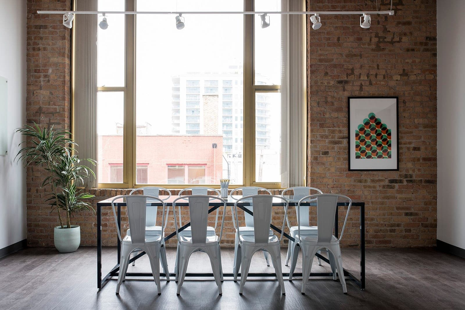 A small meeting room with a brick wall, a tall window, a long table and white metal chairs