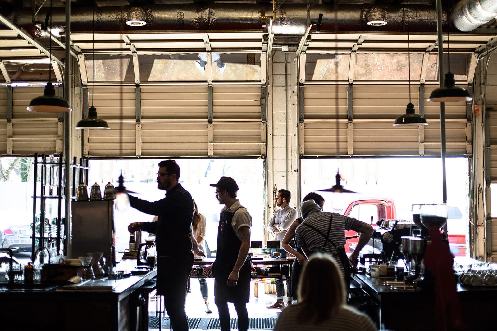 Staff work behind a long coffee bar in a busy café with open garage-door windows, seen against the daylight