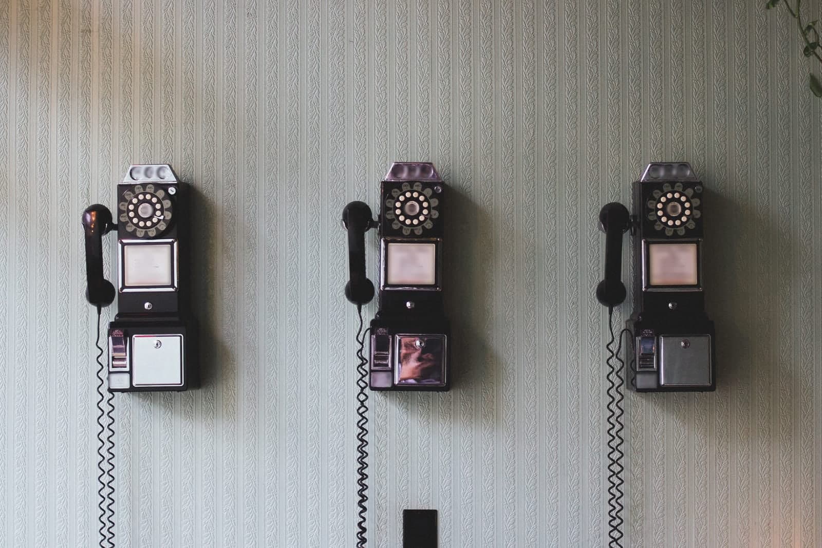 Three old-style telephones with coiled cords mounted in a row on a pale patterned wall