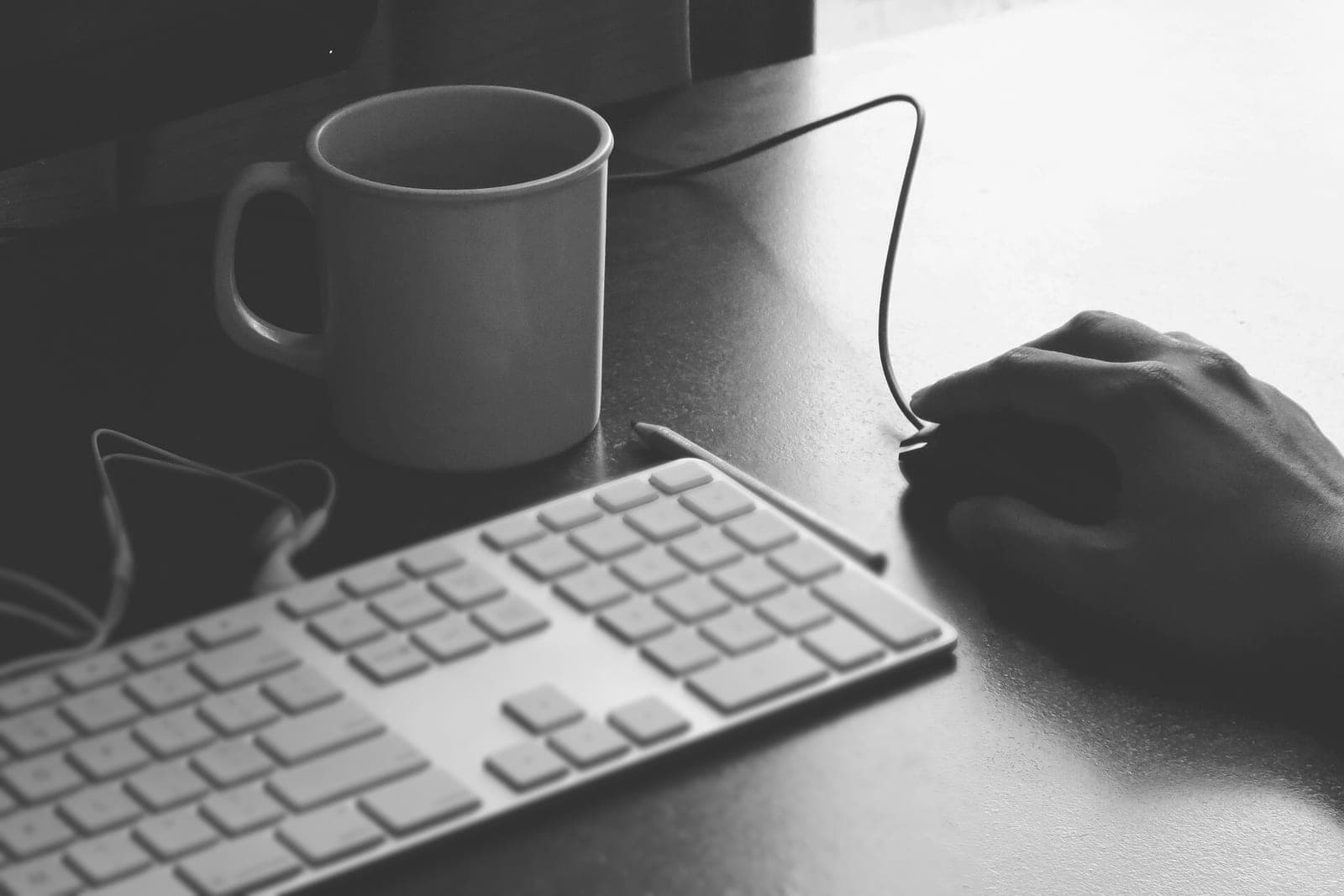A hand rests on a computer mouse beside a keyboard and a coffee mug in black and white