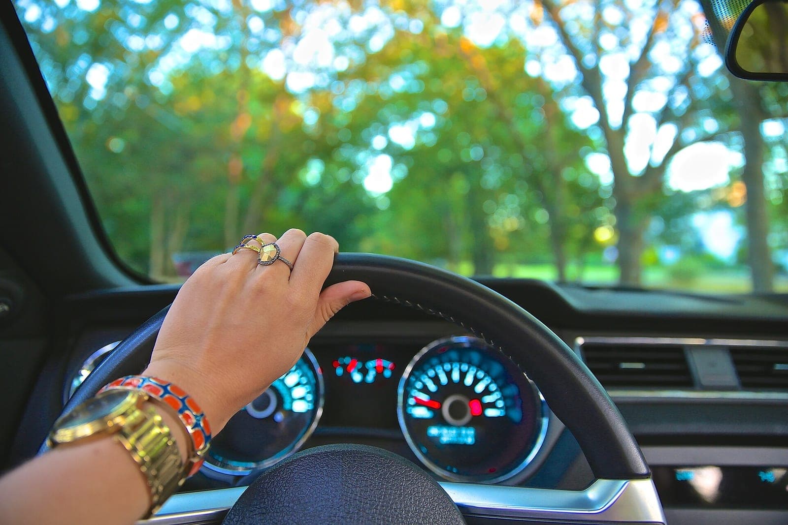 A driver's hand on a steering wheel above a lit dashboard, with green trees through the windshield