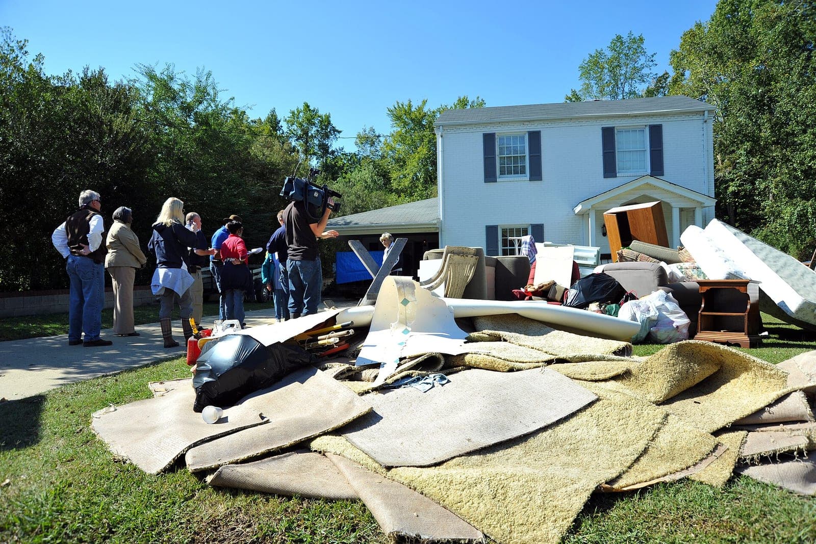Soaked carpet and furniture piled on the lawn outside a white two-story house, with a group of people standing on the driveway